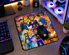 A large rectangular gaming mousepad with a colorful Yu-Gi-Oh! anime character print, shown on a wooden desk with a black gaming mouse resting on it. The desk setup includes a keyboard, monitor, PC tower with RGB lighting, and headphones.