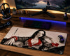 A large gaming desk mat featuring a stylized black-and-white portrait of a woman with bold red accents, shown on a wooden desk alongside a keyboard, mouse, headphones, and monitor in a lit gaming setup.