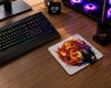 A large rectangular gaming mousepad with a colorful Yu-Gi-Oh! anime character print, shown on a wooden desk with a black gaming mouse resting on it. Surrounding the mousepad are a keyboard, monitor, PC tower with RGB lighting, and headphones.