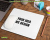 A white rectangular desk mat with rounded corners on a wooden desk, printed with the black text 'YOUR IDEA WE DESIGN'. A white computer mouse rests on the left side of the mat.