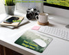 A custom desk mat with a green forest landscape print and yellow placeholder text, shown on a white desk alongside a keyboard, mouse, camera, laptop, and mug. The desk mat has a 'PokettoG' watermark overlay.