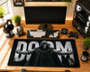 A large black gaming desk mat featuring a printed graphic of a hooded grim reaper with outstretched hands and the word 'DOOM' in large white letters, styled on a wooden desk with a keyboard, mouse, headset, and computer setup.