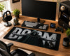 A large black gaming desk mat featuring a graphic of a hooded figure with outstretched arms and the word 'DOOM' in bold white letters, styled on a wooden desk with a keyboard, monitor, headphones, and other desk accessories.