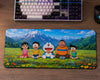 A large rectangular desk mat featuring an illustrated scene of Doraemon characters sitting in a flower field with mountains in the background, placed on a wooden desk below a keyboard and next to a mouse.