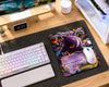 A gaming desk setup featuring a large dark gray mousepad with a colorful Pokémon trading card design, a white Logitech keyboard, a white Logitech mouse, and a monitor on a light wood desk.