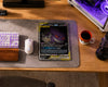 A Pokémon Trading Card Game card featuring Mirror Moon GX is displayed on a gray desk mat on a wooden desk, surrounded by a keyboard, mouse, and a Pokémon Ball mug.