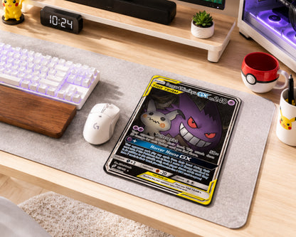 A Pokémon trading card featuring Ho-Oh GX displayed on a gray desk mat on a wooden desk, surrounded by a keyboard, mouse, mug, and other desk accessories.