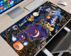 A large desk mat featuring a detailed Pokémon illustration with Gengar, Togepi, and other ghost-type creatures in a spooky night scene, shown on a light wood desk with a keyboard, mouse, and monitor nearby.
