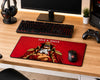 A large gaming desk mat featuring an anime-style illustration of Gold Roger from One Piece on a red background, styled on a wooden desk alongside a keyboard, mouse, headphones, and controller.