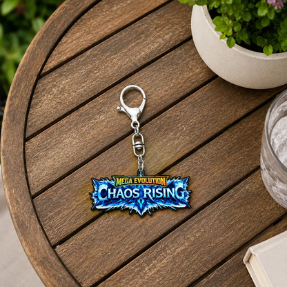 A MEGA EVOLUTION Chaos Rising keychain with a silver clasp, resting on a round wooden table next to a potted plant and a glass of water.
