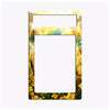 A decorative picture frame or magnet backing with a colorful landscape border. The top section features a bright yellow and white sky with sun rays, while the bottom is decorated with yellow flowers and green leaves.