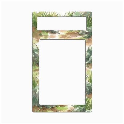 A rectangular decorative frame or magnet with a watercolor-style nature border featuring green grass, gray rocks, and brown earth, with two blank white cutout areas near the top.