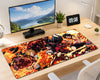 A large rectangular desk mat with a vivid anime-style flame battle scene, displayed on a light wood desk next to a computer monitor, small potted plants, a digital clock, and a desk lamp.