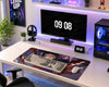 A modern gaming desk setup featuring a large anime-themed desk mat with a keyboard and mouse, a curved monitor, a desktop computer tower, and various desk accessories in a brightly lit room.