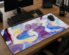 A large rectangular desk mat with a colorful anime-style character print lies on a wooden desk, surrounded by a keyboard, mouse, monitor, and mug.