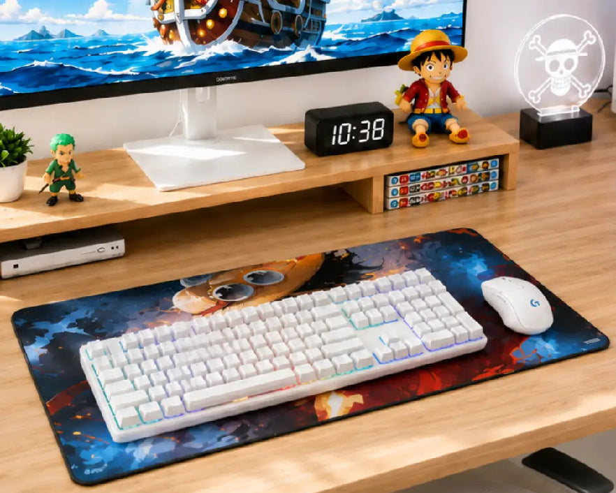 A large desk mat with a colorful cosmic and character print is shown on a wooden desk, styled with a white mechanical keyboard and a white mouse. The setup includes a monitor stand with anime figurines and a monitor in the background.