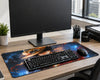 A large desk mat with a fiery cartoon character design is shown on a wooden desk, styled with a black keyboard, mouse, and a Dell monitor in a modern office setting.