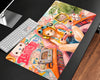 A large rectangular desk mat with a colorful anime-style illustration, shown on a dark desk next to a white keyboard and monitor stand.