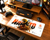 A large desk mat featuring Naruto anime artwork and the word 'NARUTO' is laid out on a wooden desk in a gaming setup, surrounded by a keyboard, mouse, controller, headphones, and monitor.