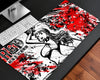 A large desk mat with a bold anime-style illustration and red, black, and white paint-splatter design, shown on a dark desk next to a keyboard and monitor stand.