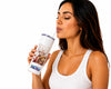 A woman with dark hair wearing a white ribbed tank top is shown from the side, drinking from a white insulated tumbler with a clear straw. The tumbler features a printed graphic of a white rose and a brown heart, with a blue and white patterned band at the bottom.