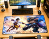 A large rectangular gaming desk mat featuring a detailed anime battle scene with two characters, placed on a wooden desk in front of a keyboard, mouse, speakers, and monitor.