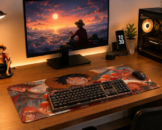 A large desk mat featuring a colorful One Piece anime illustration with Luffy, laid out on a wooden desk in a gaming setup alongside a keyboard, mouse, computer monitor, and PC tower.
