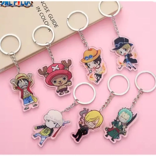 A set of seven anime keychains featuring characters from One Piece, arranged on a pink background with a beige book in the corner. The keychains are made of translucent acrylic with silver metal key rings and chains.