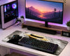 A large desk mat with a mountain and boat graphic design is shown on a white desk alongside a black keyboard, mouse, and a computer tower with a game controller. The desk setup is illuminated by purple ambient lighting.