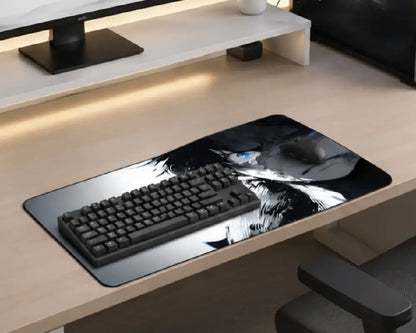 A large desk mat with a black and white anime-style graphic lies on a light wood desk, styled with a black keyboard and mouse. The desk is part of a modern home office setup with a white shelf and monitor stand in the background.