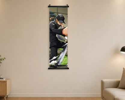 A vertical wall scroll poster hanging on a beige wall in a modern living room setting. The poster features a printed graphic of a man in a black tracksuit and cap working on a bicycle.
