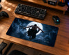 A large rectangular gaming mousepad with a dark fantasy print of a samurai floating in front of a full moon, shown on a wooden desk alongside a keyboard, mouse, and controller.