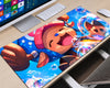 A large desk mat featuring colorful anime-style artwork of a character surrounded by bubbles, shown on a light wood desk next to a computer monitor, keyboard, and mouse.