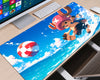 A large desk mat featuring a colorful anime-style print of a character flying over blue ocean waves with a red-and-white striped ball, shown on a wooden desk next to a computer monitor, keyboard, and mouse.