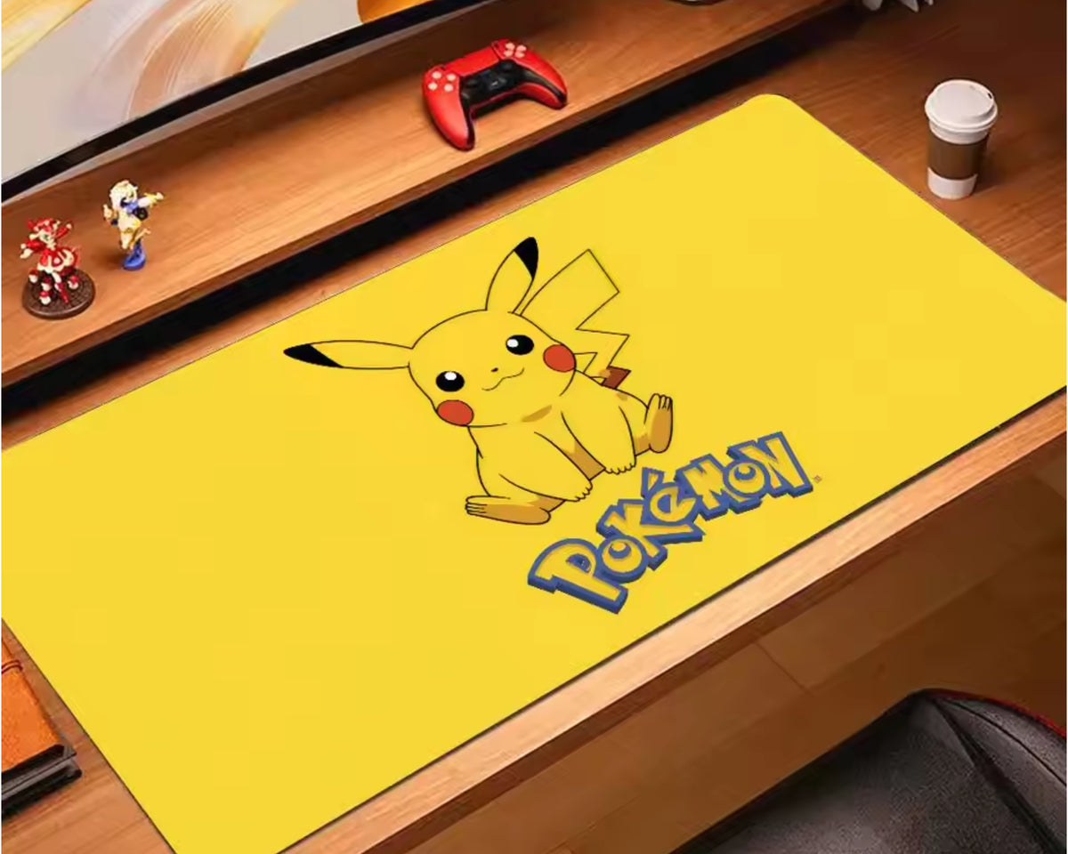 Bright yellow Pokémon extended gaming mouse pad featuring a cute Pikachu illustration with black-tipped ears, red cheeks, and the Pokémon logo, displayed on a white computer desk beneath a monitor with headphones and a keyboard nearby.