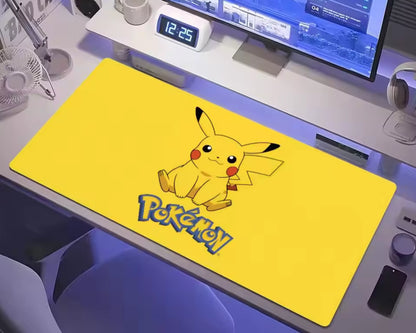 Bright yellow Pokémon extended gaming mouse pad featuring a cute Pikachu illustration with black-tipped ears, red cheeks, and the Pokémon logo, displayed on a white computer desk beneath a monitor with headphones and a keyboard nearby.