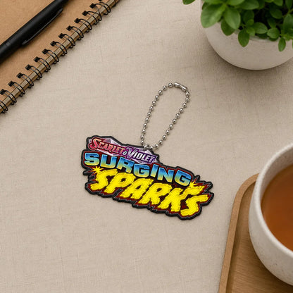 A 'Scarlet Voltage: Surging Sparks' keychain or bag charm with a silver ball chain, styled on a beige textured desk with a notebook, plant, and cup of tea.