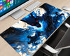 A large desk mat featuring a dynamic blue and black anime-style illustration of a dragon and warrior, shown on a light wood desk next to a computer monitor, keyboard, mouse, and office chair.