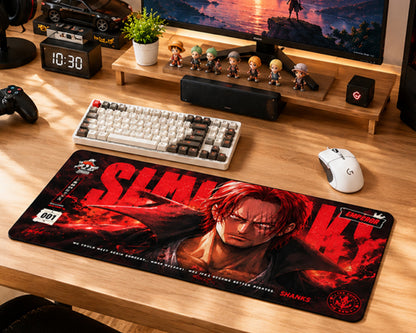 A large rectangular desk mat featuring a stylized anime character print with the name 'Shanks' and red graphic text, placed on a wooden desk in a gaming setup. The desk mat is surrounded by a keyboard, mouse, controller, speaker, and other desk accessories.