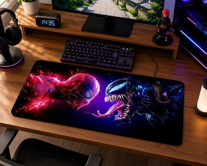 A large gaming desk mat featuring a dramatic Spider-Man and Venom face-off design, shown on a wooden desk alongside a keyboard, mouse, headphones, and monitor.