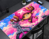 A large rectangular desk mat featuring a colorful anime-style illustration of a character with a pink hat and bicycle against a pink and blue sky background, displayed on a dark gray desk.