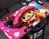 A large rectangular desk mat featuring a colorful anime-style character illustration, shown on a dark desk with a chair, phone, and small plant nearby.