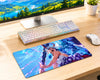 A large rectangular gaming desk mat featuring a colorful anime-style illustration of a female warrior with blue hair and lightning effects, placed on a light wood desk next to a keyboard, mouse, and computer monitor.