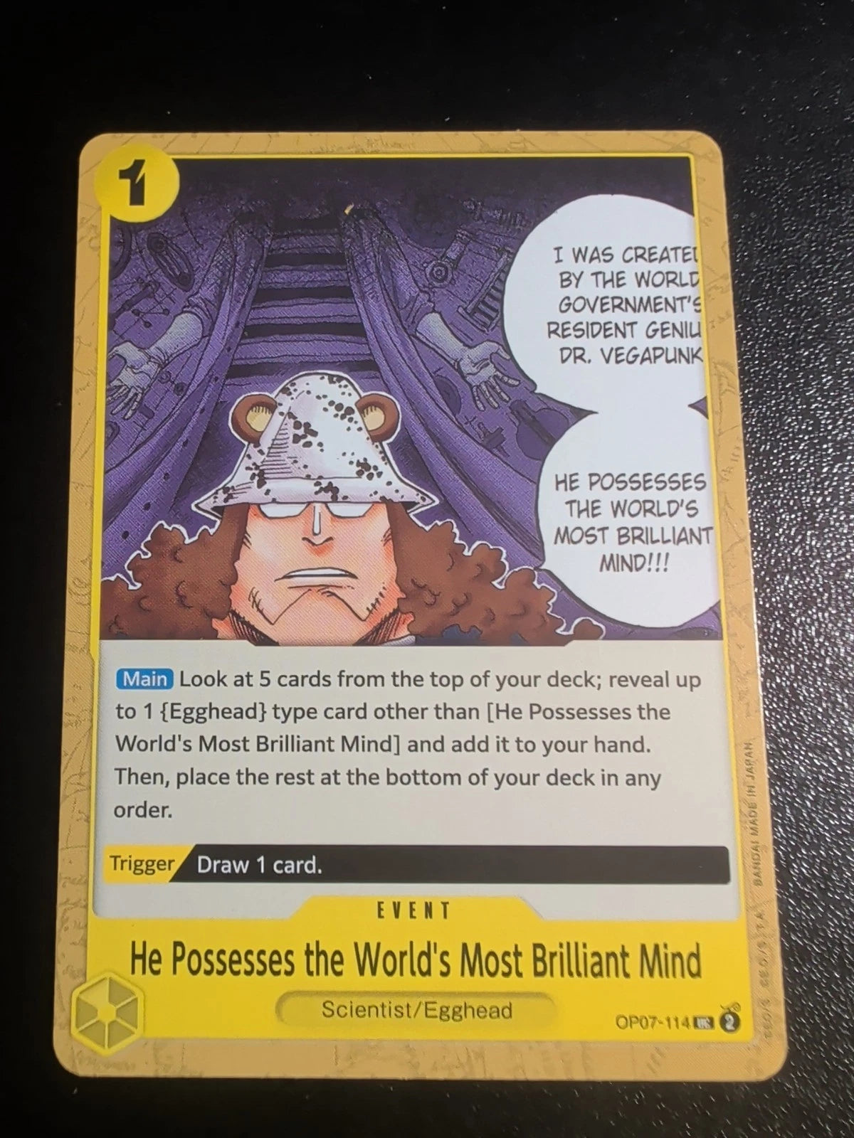 One Piece TCG event card OP07-114 featuring Bartholomew Kuma in his signature spotted hat with two speech bubbles describing Dr. Vegapunk's brilliant mind.